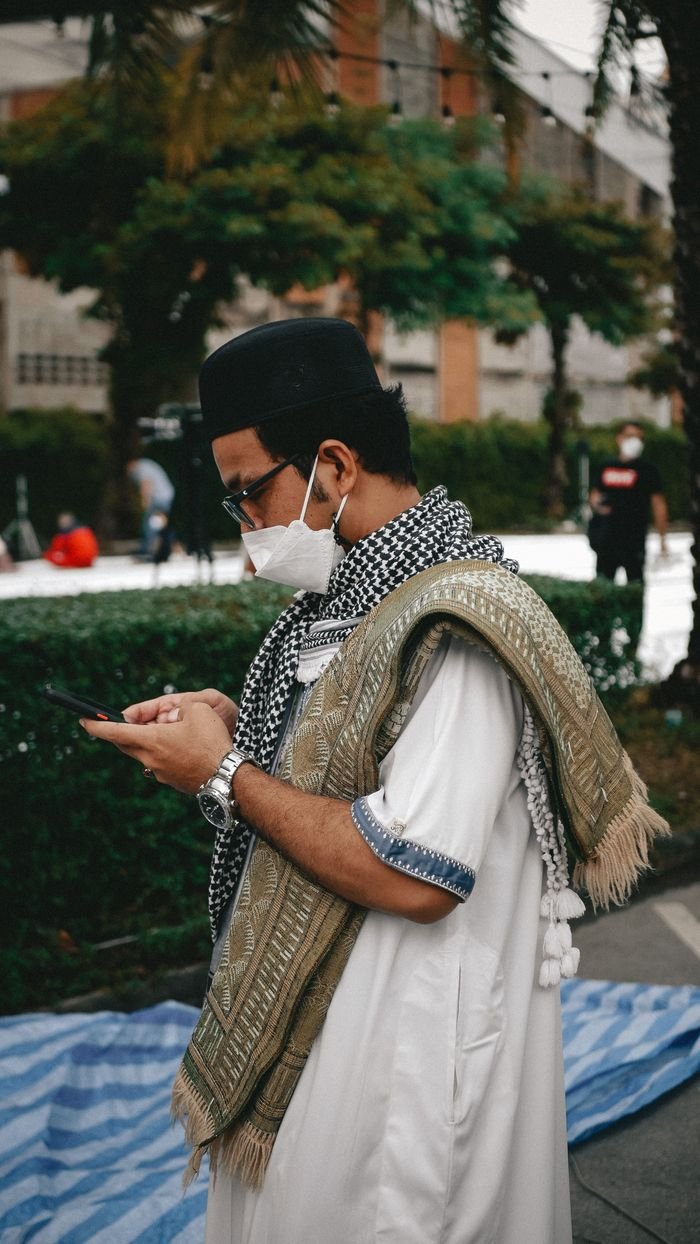 A Muslim man wearing traditional attire and facemask using a smartphone outdoors during the pandemic.