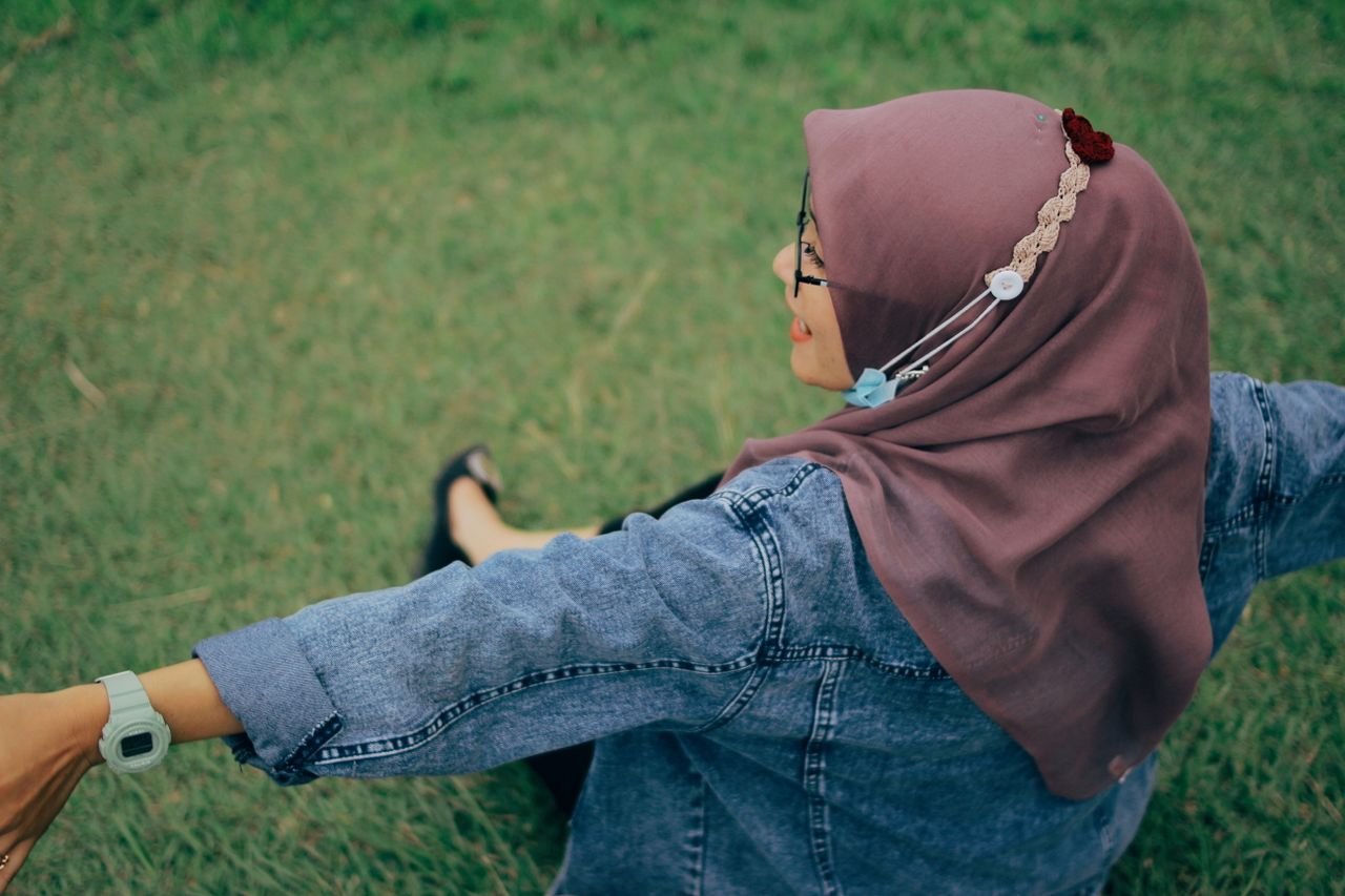 Woman wearing a hijab and denim jacket sitting outdoors on grass, enjoying a peaceful day.